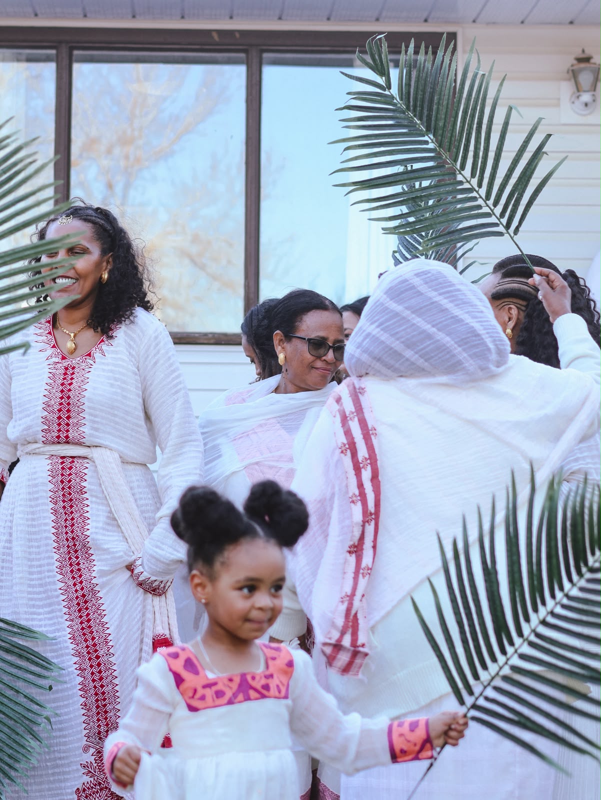 Woman in a red-embroidered white dress smiling, with family and children gathered under palm branches
