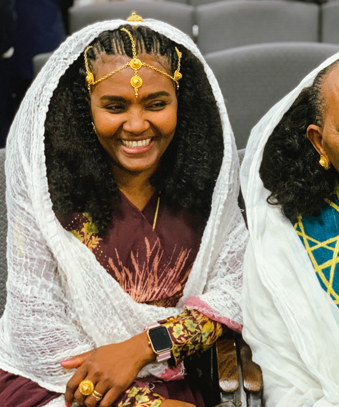 Woman smiling in a headscarf with gold jewelry, seated beside a friend